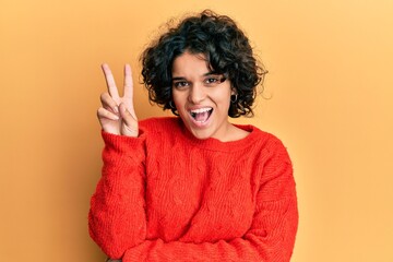 Young hispanic woman with curly hair wearing casual winter sweater smiling with happy face winking at the camera doing victory sign with fingers. number two.