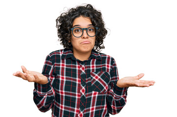 Young hispanic woman with curly hair wearing casual clothes and glasses clueless and confused...