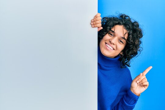 Young hispanic woman with curly hair holding blank empty banner smiling happy pointing with hand and finger to the side