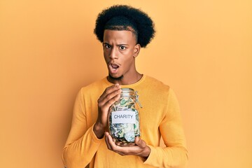 African american man with afro hair holding charity jar with money in shock face, looking skeptical...