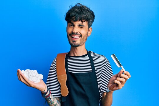 Young hispanic man wearing barber apron holding razor and foam winking looking at the camera with sexy expression, cheerful and happy face.