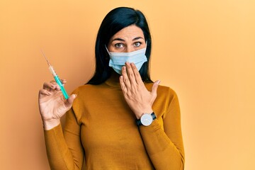 Young caucasian woman wearing medical mask holding syringe covering mouth with hand, shocked and afraid for mistake. surprised expression