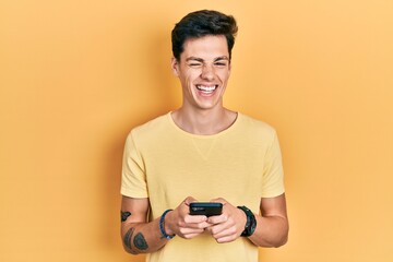 Young hispanic man using smartphone typing message winking looking at the camera with sexy expression, cheerful and happy face.