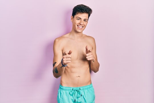 Young hispanic man wearing swimwear shirtless pointing fingers to camera with happy and funny face. good energy and vibes.