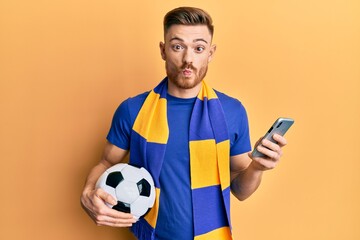 Young redhead man wearing hooligan scarf and using smartphone making fish face with mouth and squinting eyes, crazy and comical.