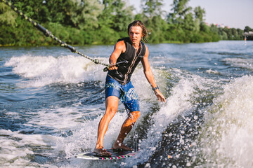 Fototapeta premium Young athletic man with long hair wakesurfing on waves of river in sunny summer weather. Ttheme outdoor activities in summer. Water sports wakesurf on the board. sliding wakeboarder in water splash