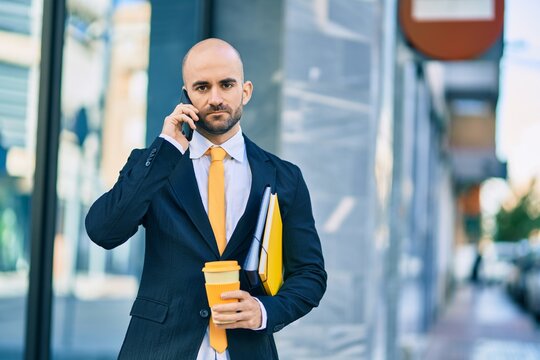 Young hispanic bald businessman with serious expression talking on the smartphone drinking coffee at the city