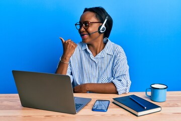 Young african woman wearing call center agent headset pointing thumb up to the side smiling happy with open mouth