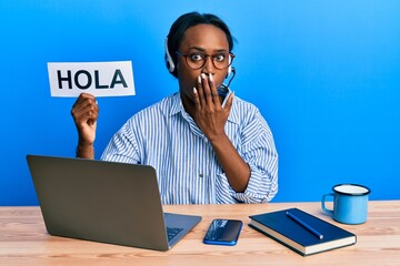 Young african woman wearing operator headset holding hola word covering mouth with hand, shocked and afraid for mistake. surprised expression © Krakenimages.com