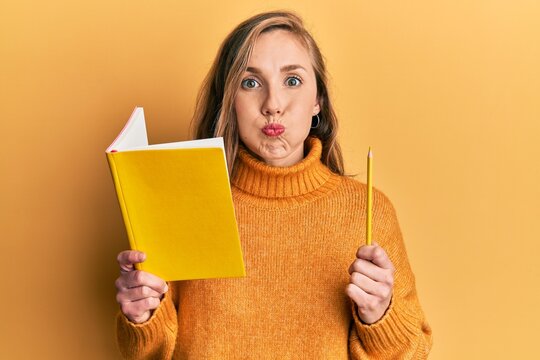 Young blonde woman holding book and pencil puffing cheeks with funny face. mouth inflated with air, catching air.