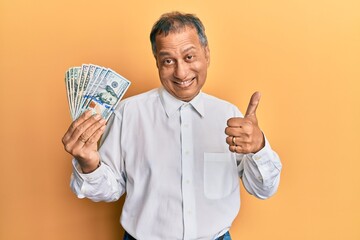 Middle age indian man holding dollars smiling happy and positive, thumb up doing excellent and approval sign