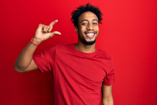 Young african american man with beard wearing casual red t shirt smiling and confident gesturing with hand doing small size sign with fingers looking and the camera. measure concept.