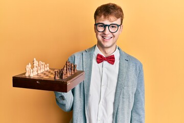 Young caucasian man wearing glasses holding chess board looking positive and happy standing and...