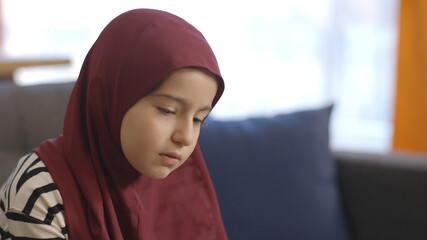 The Muslim little girl wearing a headscarf contemplates out of sorrow, holding her head in her...