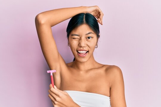 Young chinese woman wearing shower towel using razor winking looking at the camera with sexy expression, cheerful and happy face.