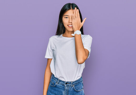 Beautiful young asian woman wearing casual white t shirt covering one eye with hand, confident smile on face and surprise emotion.