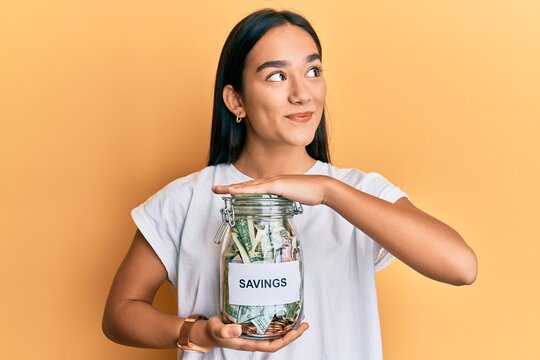 Young asian woman holding jar with savings smiling looking to the side and staring away thinking.