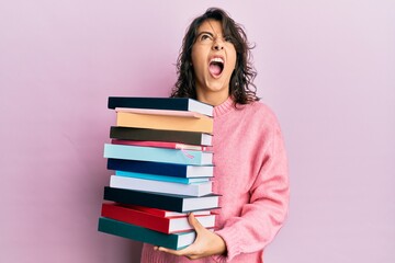 Young hispanic woman holding a pile of books angry and mad screaming frustrated and furious, shouting with anger looking up.
