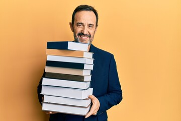 Middle age hispanic man holding a pile of books looking positive and happy standing and smiling with a confident smile showing teeth