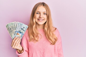 Beautiful young caucasian girl holding dollars looking positive and happy standing and smiling with a confident smile showing teeth