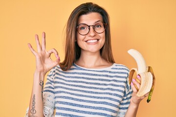 Beautiful caucasian woman eating banana as healthy snack doing ok sign with fingers, smiling friendly gesturing excellent symbol