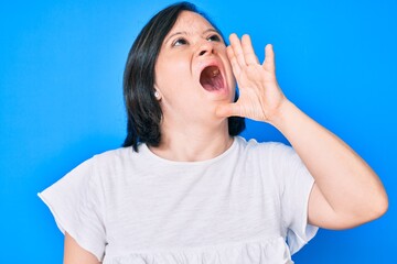 Brunette woman with down syndrome wearing casual clothes shouting and screaming loud to side with hand on mouth. communication concept.