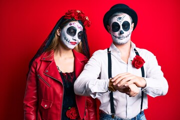 Couple wearing day of the dead costume over red checking the time on wrist watch, relaxed and confident