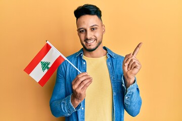 Young arab man holding lebanon flag smiling happy pointing with hand and finger to the side