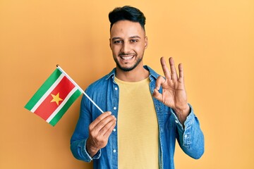 Young arab man holding suriname flag doing ok sign with fingers, smiling friendly gesturing...