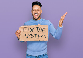 Young arab man holding fix the system banner cardboard celebrating victory with happy smile and winner expression with raised hands