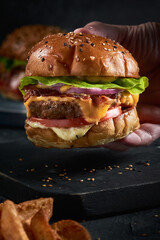 mano de hombre colocando pan a una hamburguesa doble carne con tocineta queso y cebolla frita en un fondo negro 