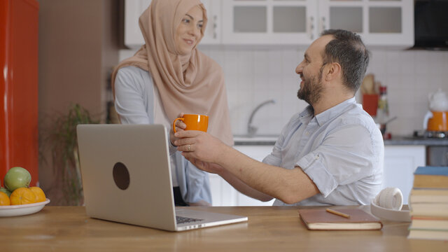 Young Muslim businessman working at home with computer during the epidemic. His wife in a turban is giving coffee to the young businessman. Home working concept. 
