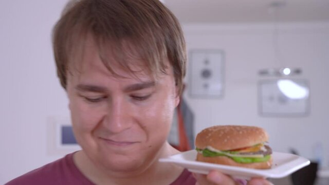 Thoughtful young man holds small plate with delicious fresh hamburger, touches lips hesitating in contemporary kitchen closeup