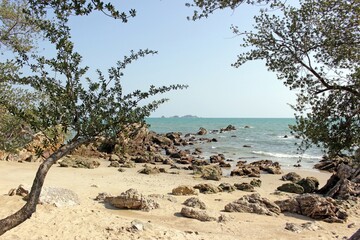 View through trees to a quiet empty tropical beach scene on a summer's day with sand and rocks going out into the ocean, southeast Asia