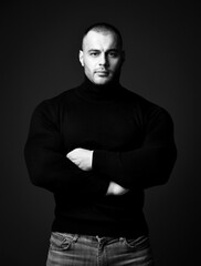 Naklejka na ściany i meble Self-confident strong man, sportsman, fitness trainer in black sweater turtleneck and jeans stands with arms crossed at chest and looking at camera on dark background