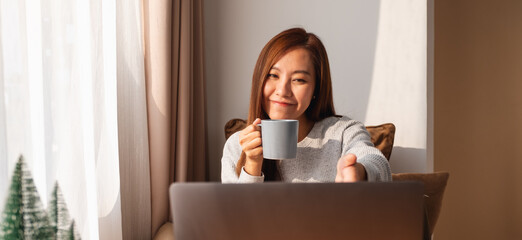 A beautiful young asian woman using laptop computer for video call while sitting and drinking coffee at home