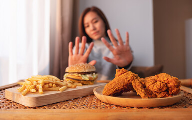 A woman making hand sign to refuse a hamburger, french fries and fried chicken on the table for...