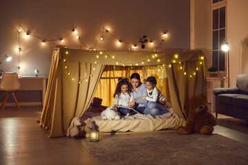 Mother with two daughters reading book and telling story hiding in tent. Happy family having fun...