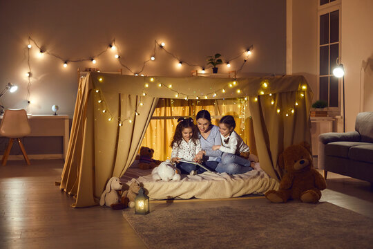 Mother with two daughters reading book and telling story hiding in tent. Happy family having fun together before going to bed in dark evening. Home living room with flashlight decoration