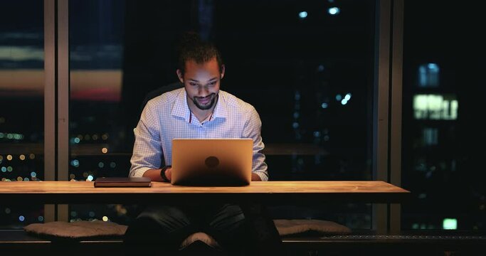 Happy young african american businessman working late on computer, sitting at table in modern luxury office with night city view, enjoying developing project or solving problems online alone indoors.
