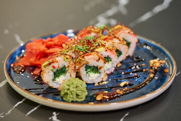 A close-up photo of sushi rolls with wakame seaweed, Philadelphia cheese, and shrimps on a blue ceramic plate with sauce, peanuts, ginger, and wasabi. Black stone table.