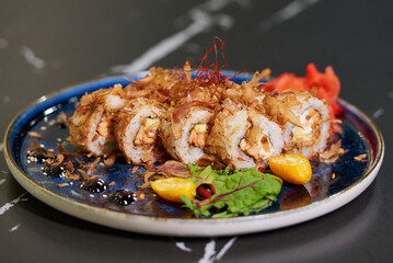A close-up photo of sushi rolls with tuna shavings, Philadelphia cheese, and eel on a blue ceramic plate with sauce, lobules of kumquat, and leaves. Black stone table.