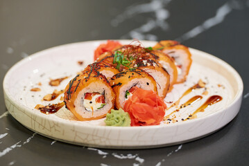 A close-up photo of sushi rolls with salmon, Philadelphia cheese, and tuna on a white ceramic plate with sauce, peanuts, ginger, and wasabi on a black stone table.