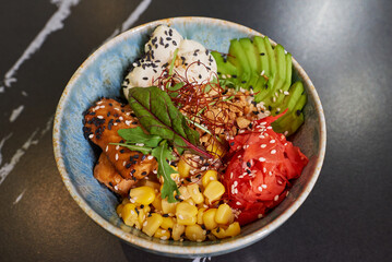 A close-up photo of a poke bowl with tuna, avocado slices, wasabi, corn, peanut, sesame, and Philadelphia cheese on a black stone table.