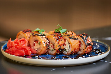 A close-up photo of fresh sushi rolls with salmon, Philadelphia cheese, and tuna on a blue ceramic plate with sauce, sesame, ginger, and wasabi. Black stone table.
