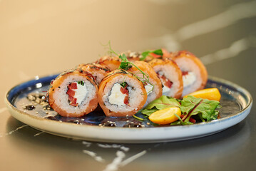 A close-up photo of fresh sushi rolls with salmon, Philadelphia cheese, and tuna on a blue ceramic plate with sauce, sesame, lobules of kumquat, and leaves. Black stone table.