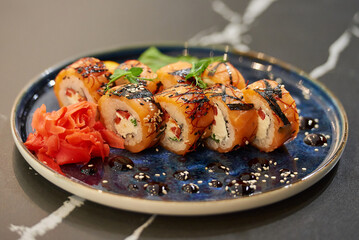 A close-up photo of sushi rolls with salmon, Philadelphia cheese, and tuna on a blue ceramic plate with sauce, sesame,  wasabi, and leaves. Black stone table.