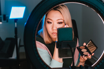 A woman of Asian appearance records video on a smartphone with a ring lamp. Girl blogger does makeup, broadcasts online. House photo