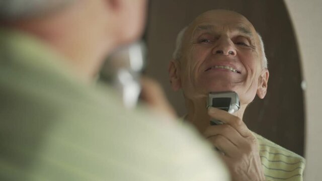 Cheerful active elderly man looking at mirror and shaving his chin with electrical shaver.