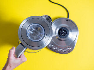 a hand holds a silver-colored electric metal kettle with different water heating modes on a yellow background. Top view, flat lay